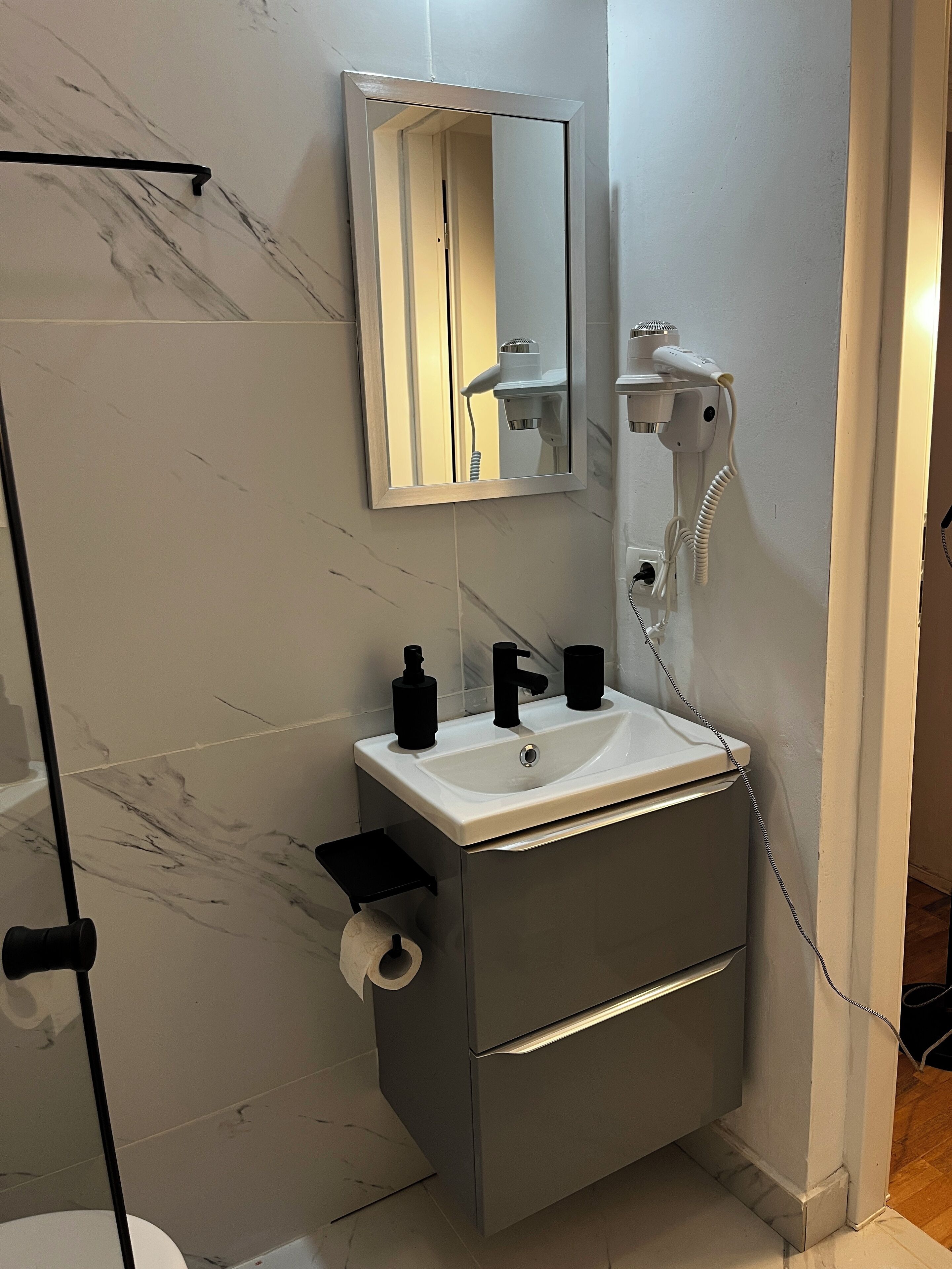 A bathroom with a white sink, black soap dispensers, a mirror, and a hairdryer mounted on the wall.