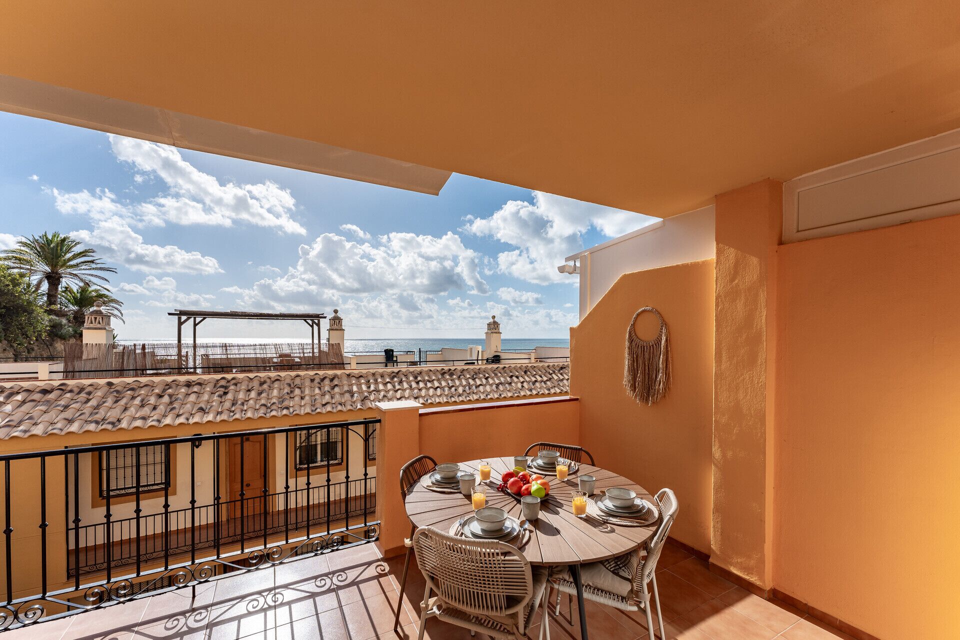 A balcony with a tiled floor, a round table set for dining, and a view of the sea and buildings in the distance.