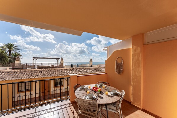 A balcony with a tiled floor, a round table set for dining, and a view of the sea and buildings in the distance.