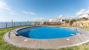 A swimming pool with a wooden deck, surrounded by a metal fence, overlooking a coastal cityscape.