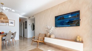 A modern living room with a large flat-screen TV mounted on the wall, a dining area with a wooden table and chairs, and a seating area with a cushioned chair and decorative vases.