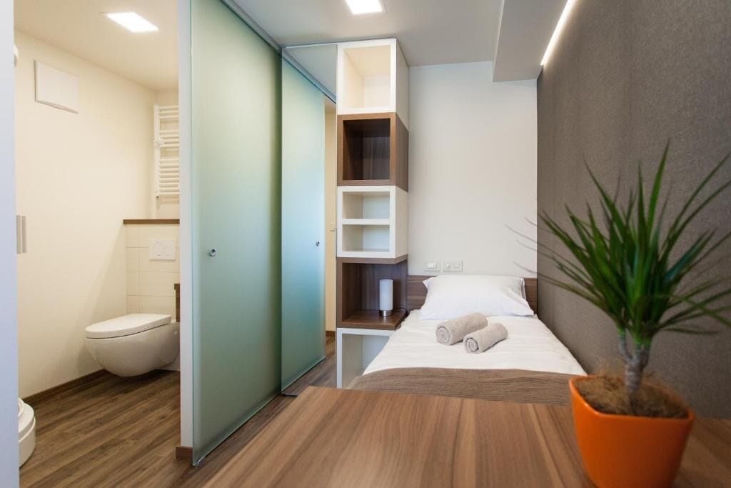 Quarto de hotel moderno com piso de madeira, cama com roupa de cama branca, mesa de cabeceira, planta em vaso e banheiro com vaso sanitário e porta-toalhas.