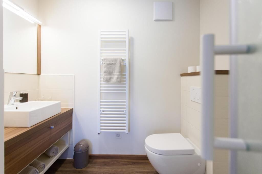 Un baño moderno con lavamanos blanco, tocador de madera, perchero, inodoro y basurero.