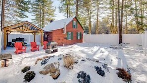 A cabin with a wooden deck, a covered area with chairs, and a snow-covered yard.