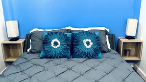 Een bed met een grijze gewatteerde spreidek en twee decoratieve kussens met een blauw-wit bloemenmotief. Aan beide zijden van het bed staan nachtkastjes met lampen.