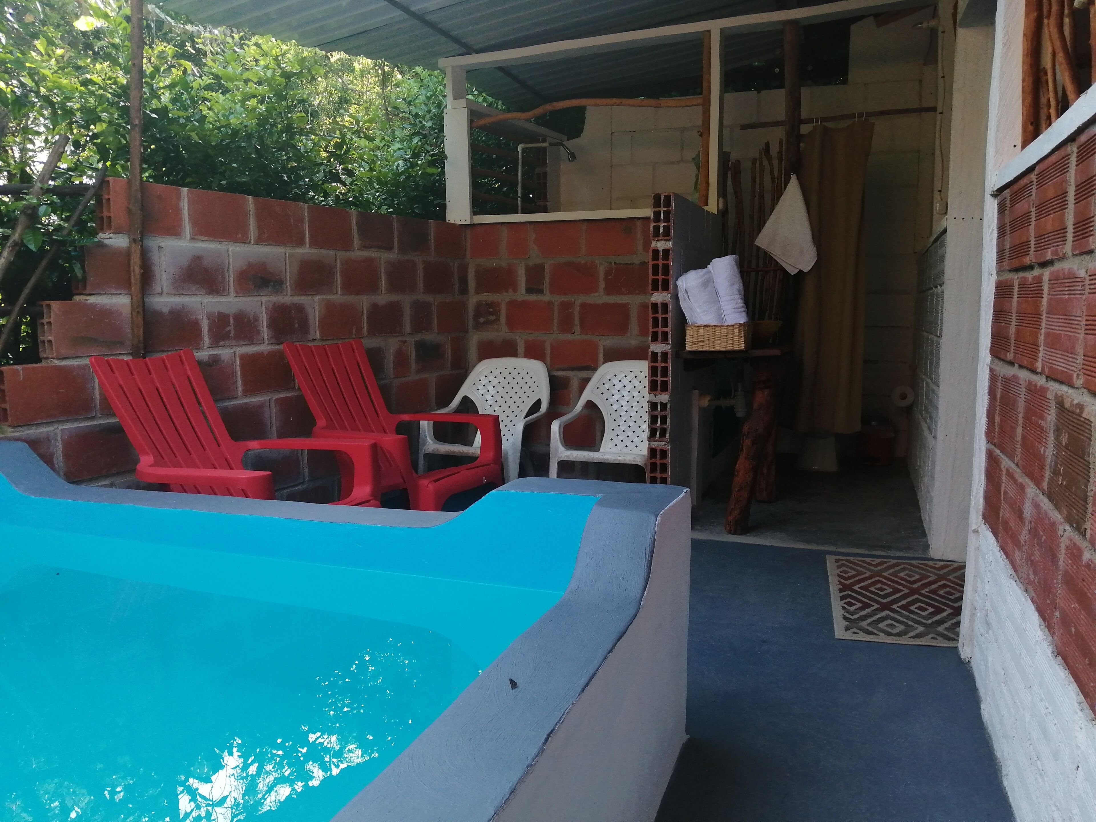 Zona de piscina con sillones reclinables rojo y blanco, un muro de ladrillo y una puerta que da a un espacio interior.