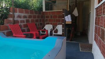 Zona de piscina con sillones reclinables rojo y blanco, un muro de ladrillo y una puerta que da a un espacio interior.