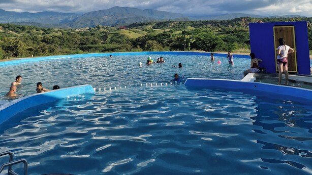 Una piscina con gente disfrutando del agua, rodeada de abundante vegetación y montañas al fondo.