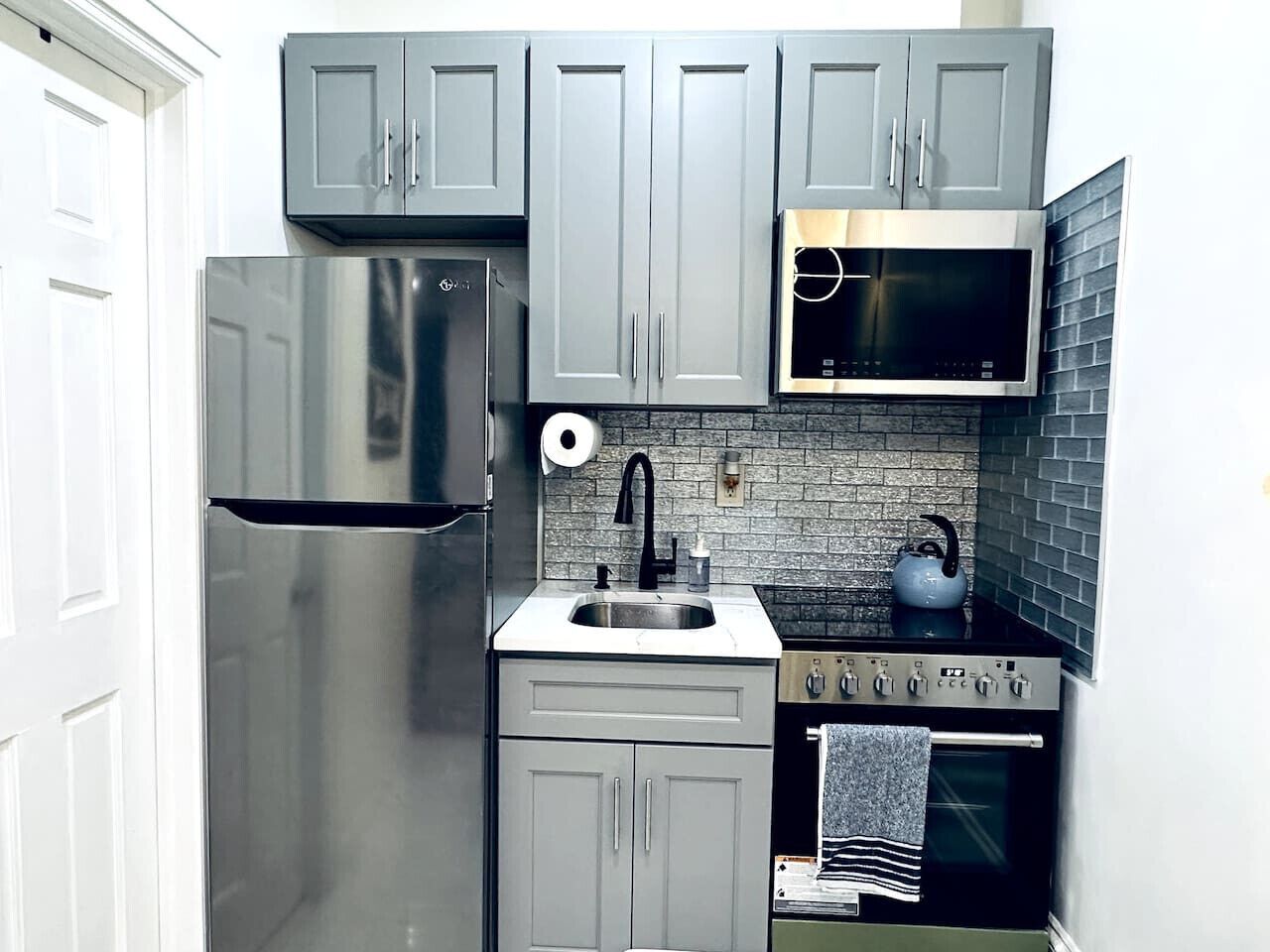 A compact kitchen with grey cabinets, a stainless steel refrigerator, a microwave, a stove, and a sink.