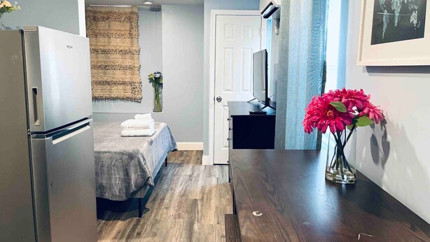 Une chambre d’hôtel de taille modeste, comprenant un lit, une petite table, un réfrigérateur, une télévision et un vase avec des fleurs.