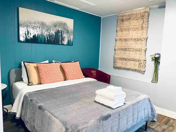 A neatly made bed with a grey blanket and multiple pillows, a wall-mounted tapestry, and a vase with flowers on a window sill. - Rittenhouse Square close by Kimmel Center Suite (Philadelphia)