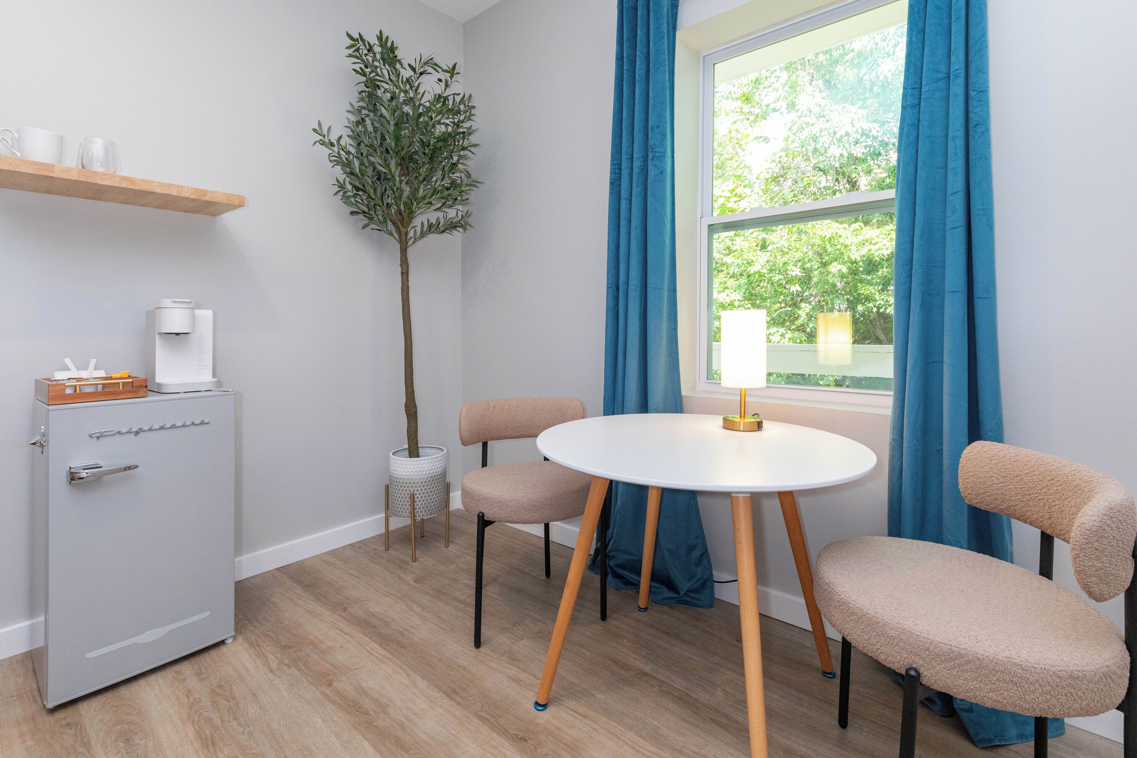 Une pièce de taille modeste comprenant un petit coin cuisine, une table ronde avec des chaises, une plante en pot et une fenêtre avec des rideaux.