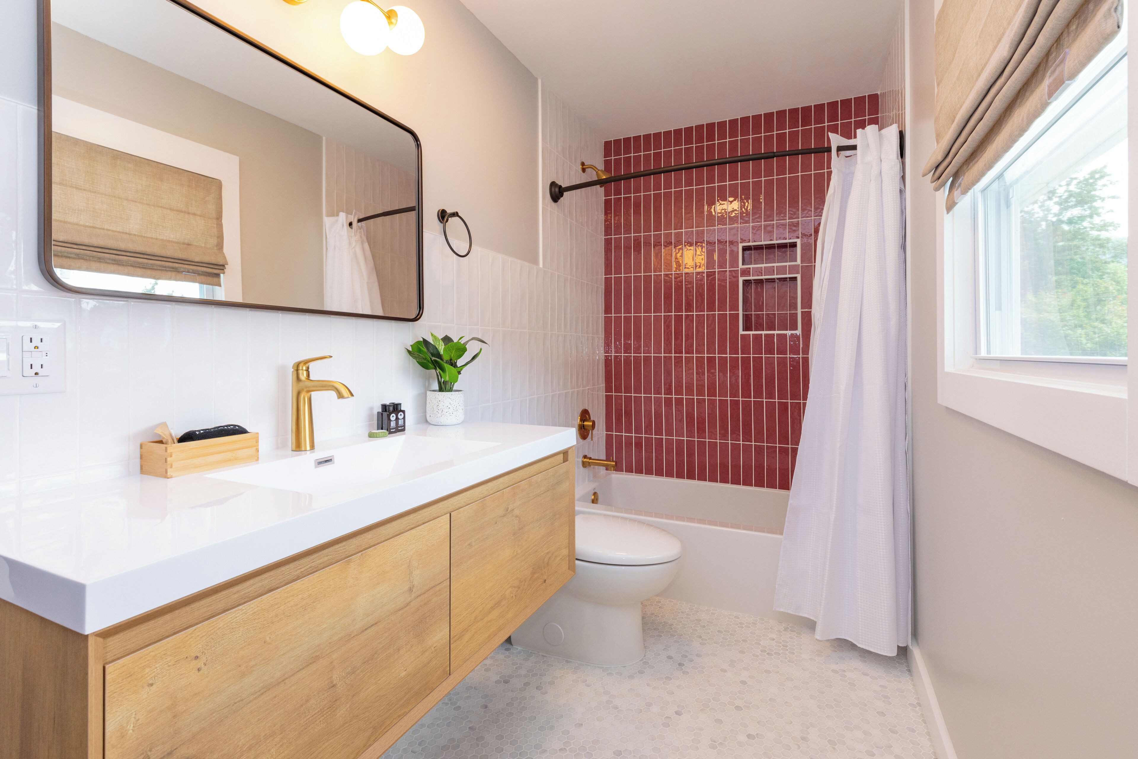 Une salle de bain moderne avec un lavabo blanc, un meuble-lavabo en bois, un grand miroir, des toilettes et une baignoire dotée d’une zone de douche carrelée en rouge.