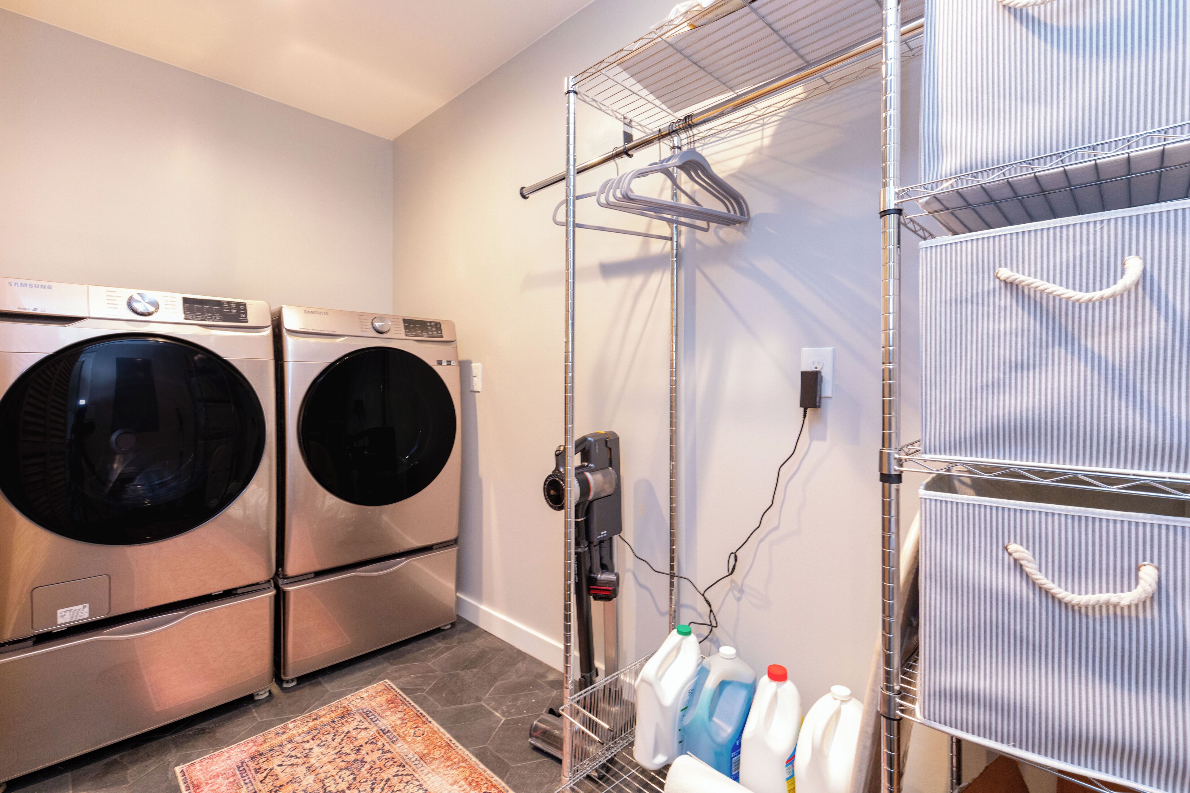A laundry room with two stacked front-loading washers and dryers, a drying rack, and shelves with detergent bottles.