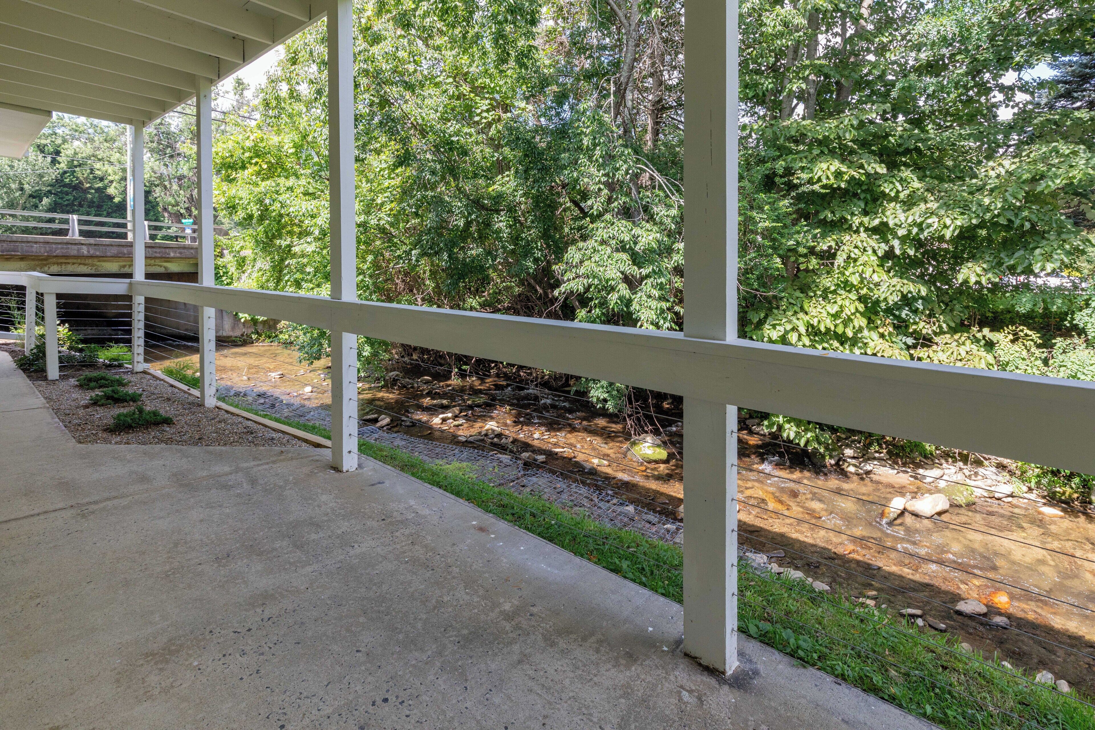 A covered outdoor area with a concrete floor, metal railings, and a view of a natural landscape with trees and a stream.
