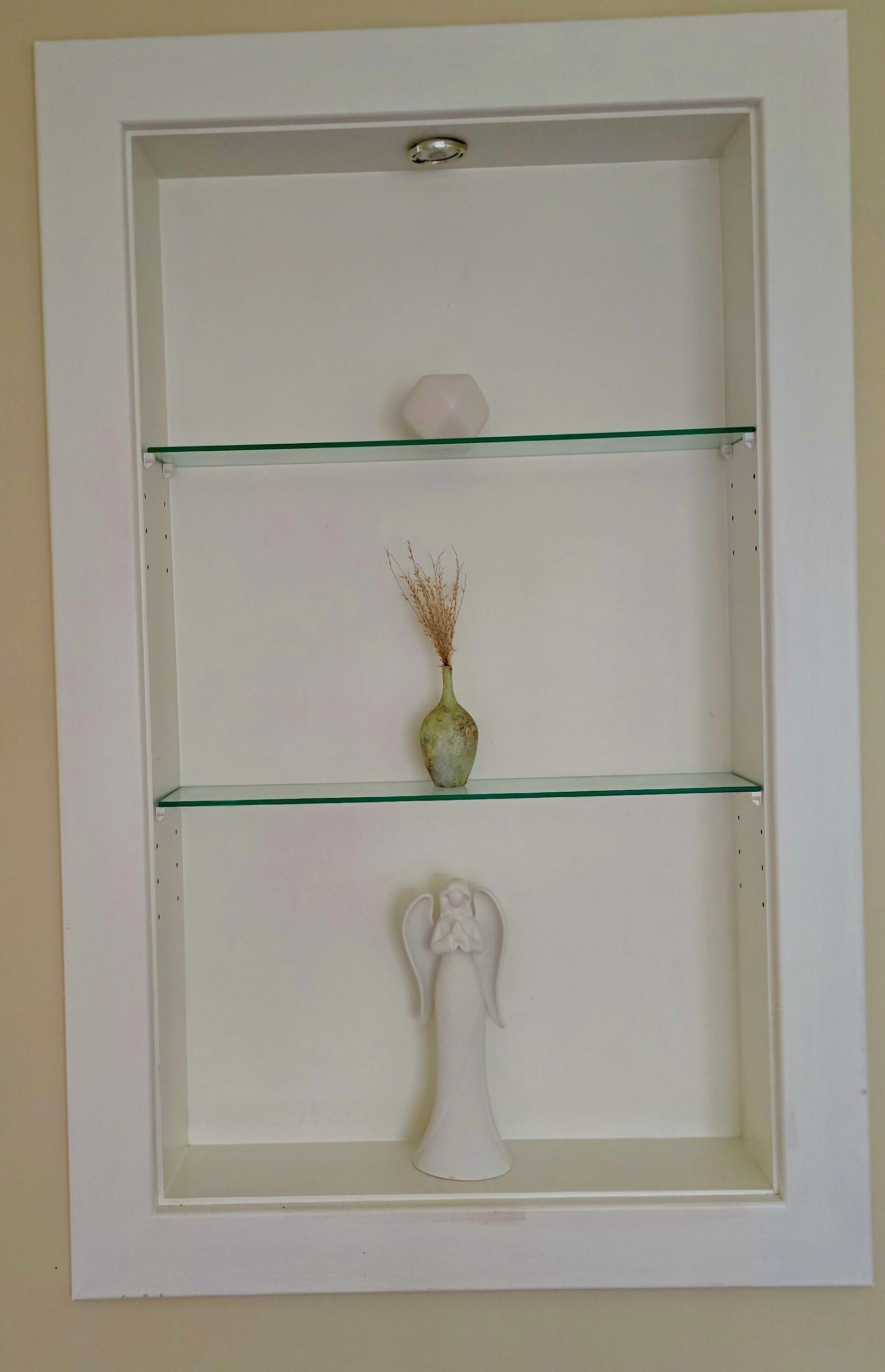 Una vetrina bianca con tre ripiani, ognuno dei quali espone un oggetto diverso: un vaso con piante essiccate, una piccola sfera e una scultura bianca.