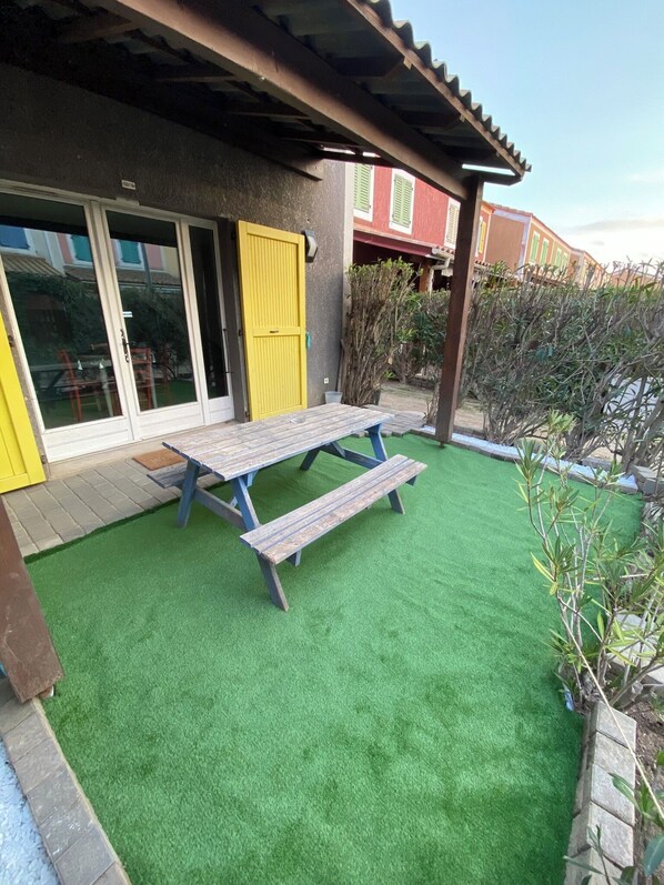 Un patio con una mesa y bancos de madera, una puerta amarilla y césped artificial verde.