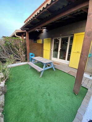Un patio con mesa y bancos de madera, una puerta amarilla y una zona cubierta con techo de tejas.