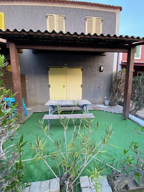 Un porche cubierto con una mesa y sillas de madera, una puerta amarilla y césped artificial verde.