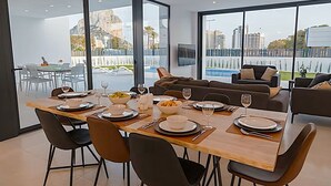 Uma sala de jantar moderna com mesa de madeira posta para quatro pessoas, grandes janelas com vista para uma piscina e prédios, e uma sala de estar com sofá e poltronas.