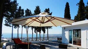 Un patio al aire libre con una mesa, sillas, un paraguas grande y vistas al mar y a la ciudad de fondo.