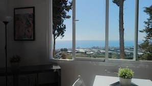 Una habitación con un ventanal grande que ofrece vistas al mar y edificios a lo lejos. Hay una mesa de comedor con una planta en maceta.