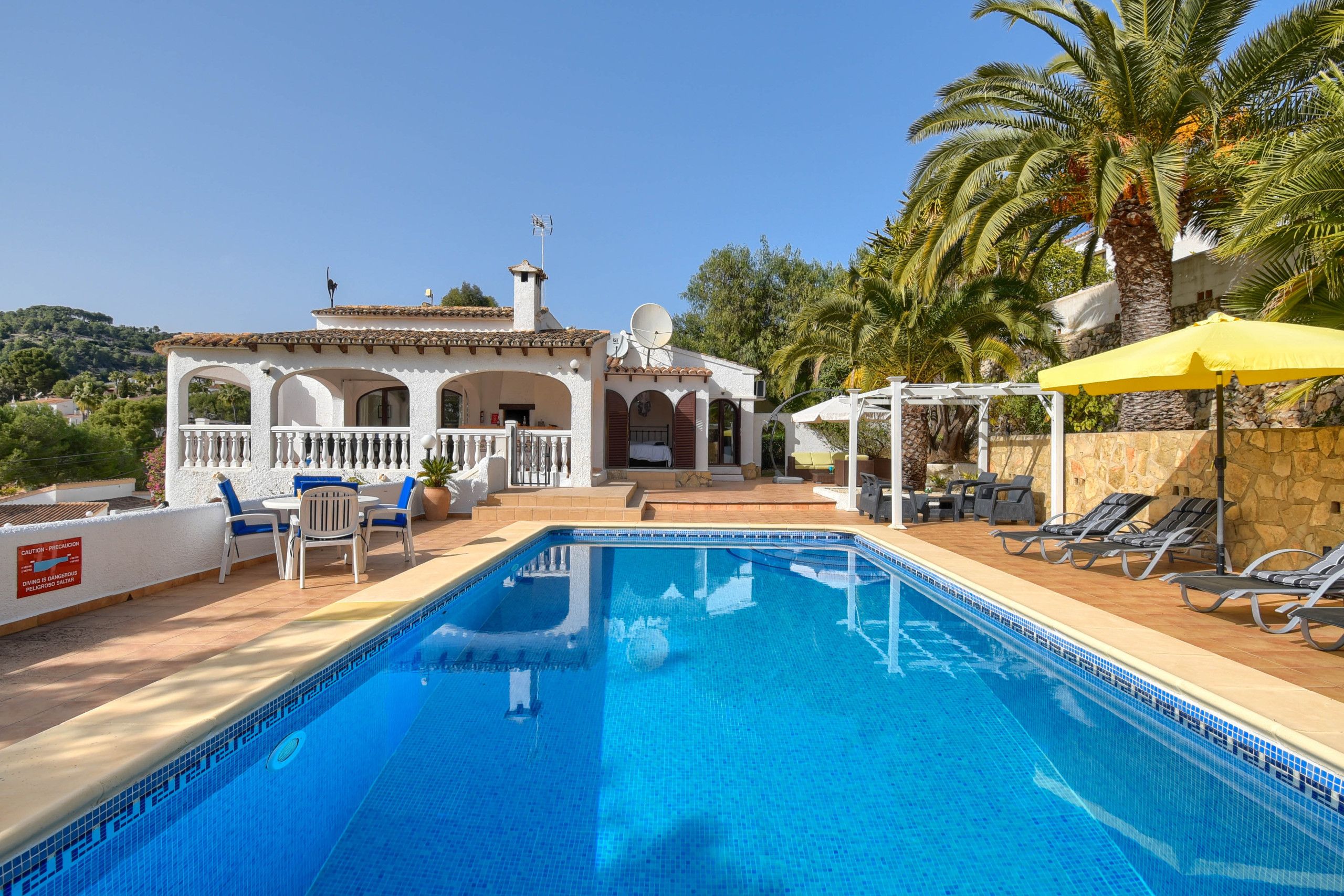 A swimming pool with lounge chairs and a patio area, surrounded by a white building with arches and a stone wall.
