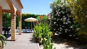 Una terraza de madera con sillones de descanso y un área de piscina, rodeada de plantas en flor y un seto.
