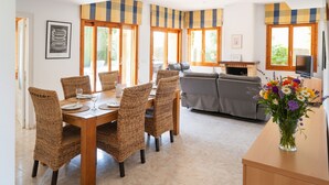 Una sala de estar moderna con zona de comedor, que incluye una gran mesa de madera, sillas de mimbre, un sofá gris y un televisor.