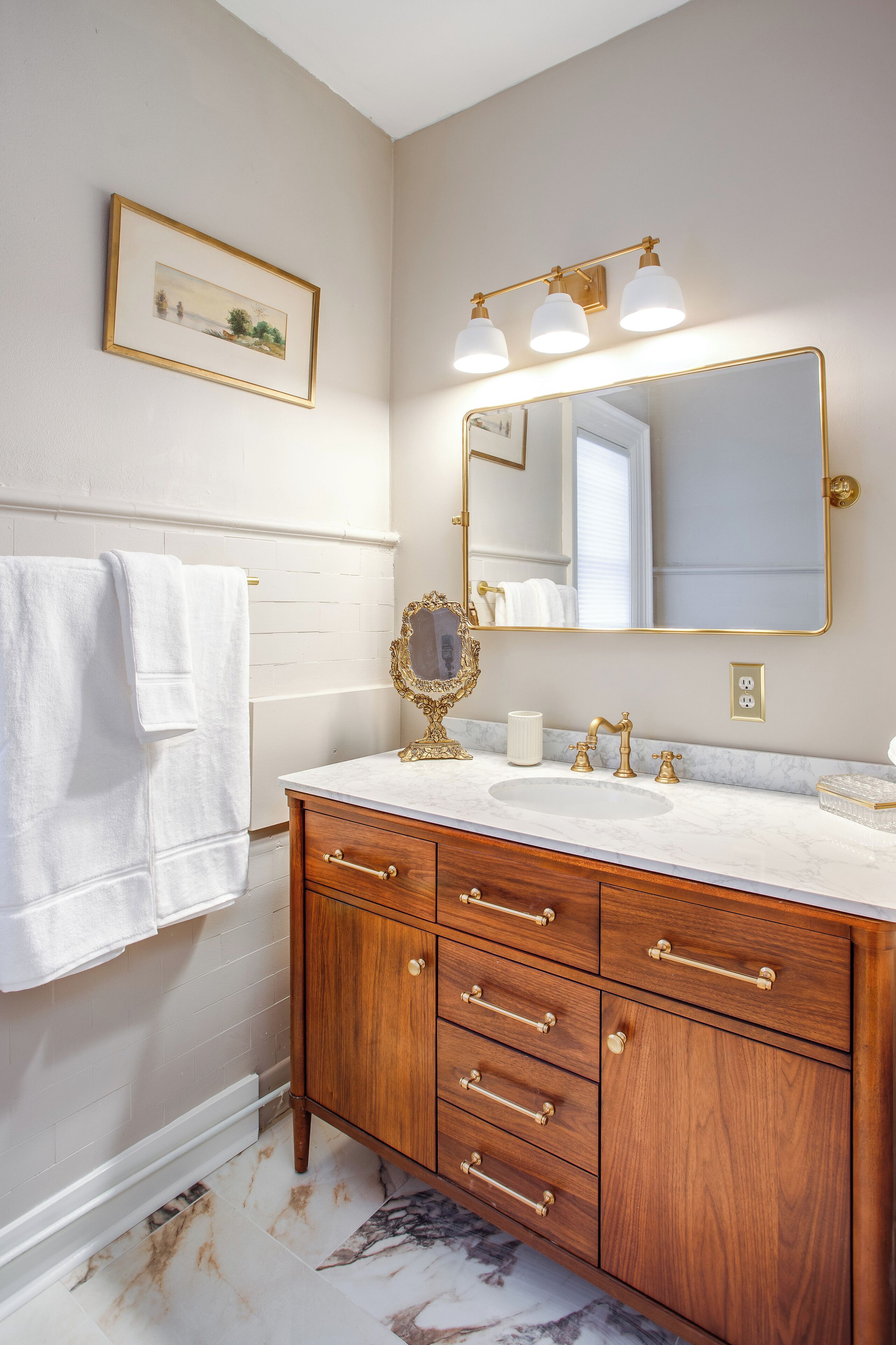 A bathroom with a marble sink, wooden vanity, gold fixtures, and a framed picture on the wall.