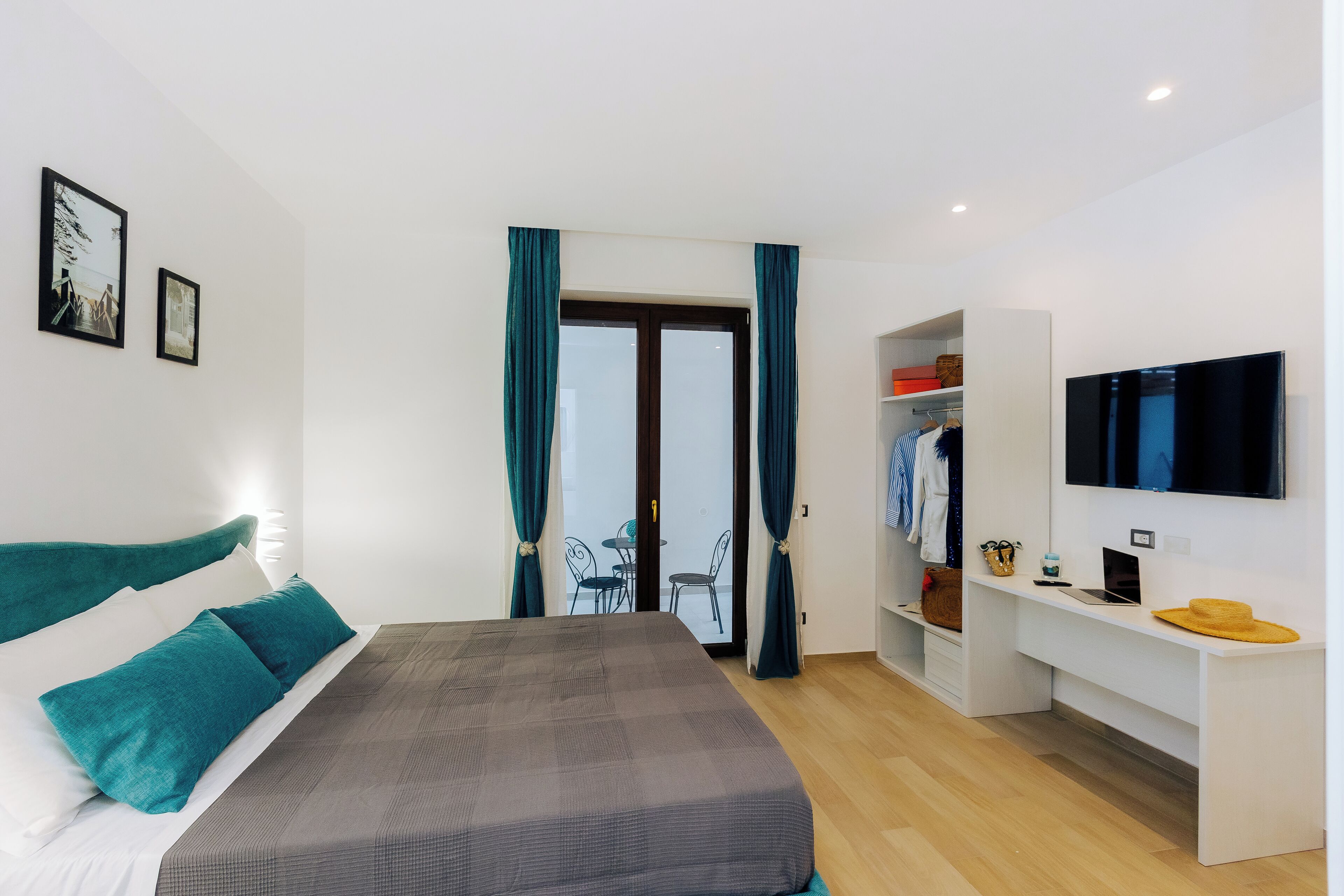 Een moderne hotelkamer met een bed, een bureau met een laptop, een televisie en een balkon met een tafel en stoelen.