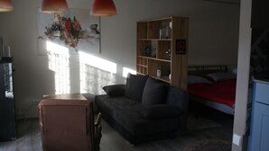 Una habitación pequeña y luminosa con un sofá, una estantería de madera, una cama con una manta roja y un cartel de 'No Smoking'.