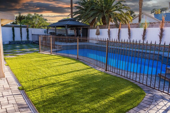 A swimming pool with a metal fence, a grassy area, and a covered seating area.