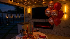 An outdoor dining area with a table set for two, featuring a charcuterie board, wine glasses, and a bottle of champagne. There are red balloons with "Happy Anniversary" written on them, and a fireplace in the background. - Beautiful and cozy family house with pool and barbecue. (Las Vegas)