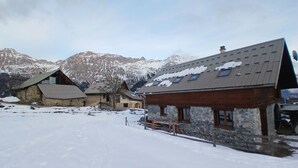 Un pueblo cubierto de nieve con edificios de piedra, una estructura de madera con techo inclinado y montañas al fondo.