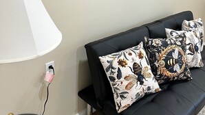 A modern living room with a black sofa, floral-patterned cushions, a large wall art piece, a white lamp, and a small table with a gold decorative item. - Huffman Extended Stay Suites (Minden)