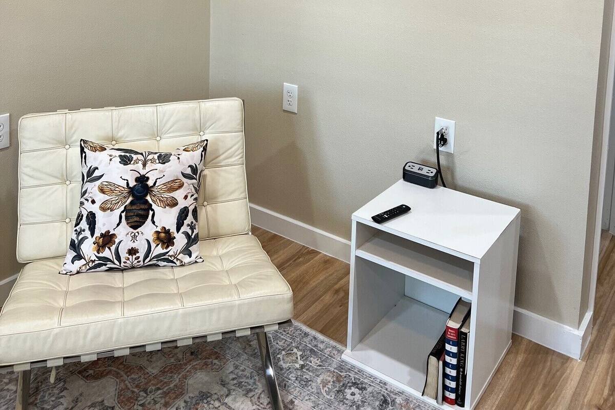 A living room with a beige sofa, a mounted TV, a small white side table with a remote, and a patterned cushion.