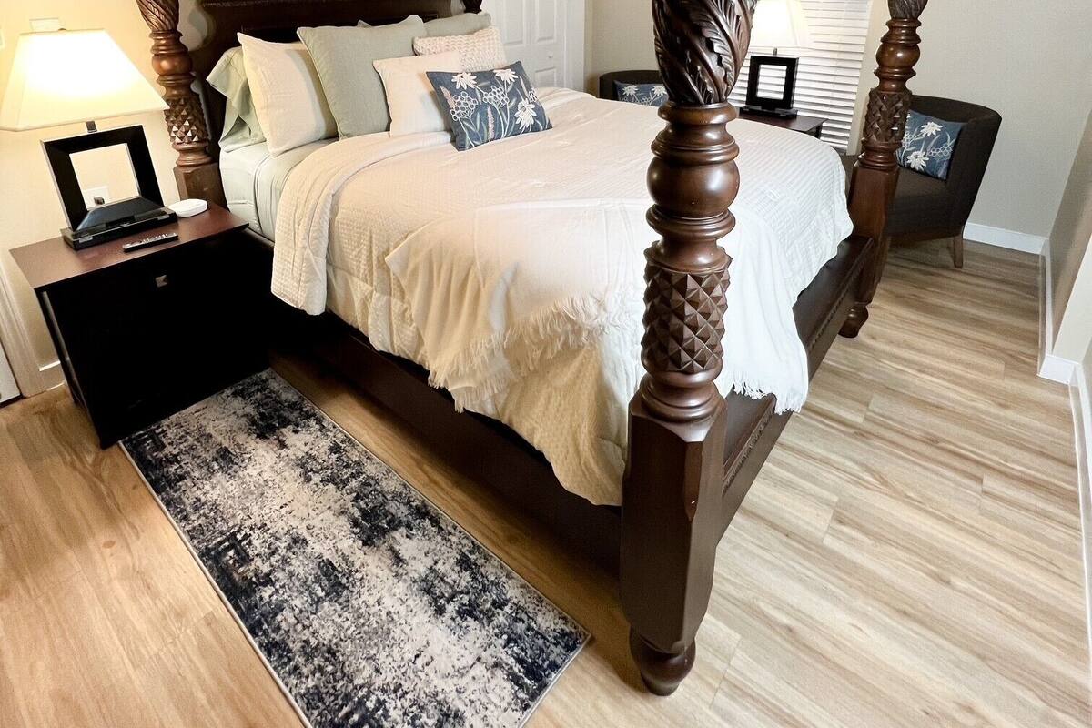 A four-poster bed with a ceiling fan, a bedside table with a lamp, and a patterned rug.