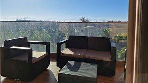 Un balcon avec deux chaises et une petite table, d’où l’on peut admirer un paysage urbain avec des bâtiments et des montagnes lointaines.