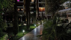 Vue nocturne d’une cour d’hôtel avec une allée pavée, illuminée par des lampes au sol, et entourée d’une végétation luxuriante et d’arbres.