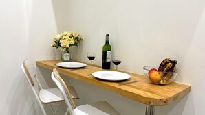 Een knus eetgedeelte met een houten tafel, twee witte stoelen, een bloevas, twee wijnglazen, een fles wijn en een schaal fruit.