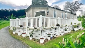 A single-story mobile home with a white exterior, a covered porch with potted plants, and a paved driveway. - Newquay Seaside Lodge Retreat With Hot Tub (Newquay)