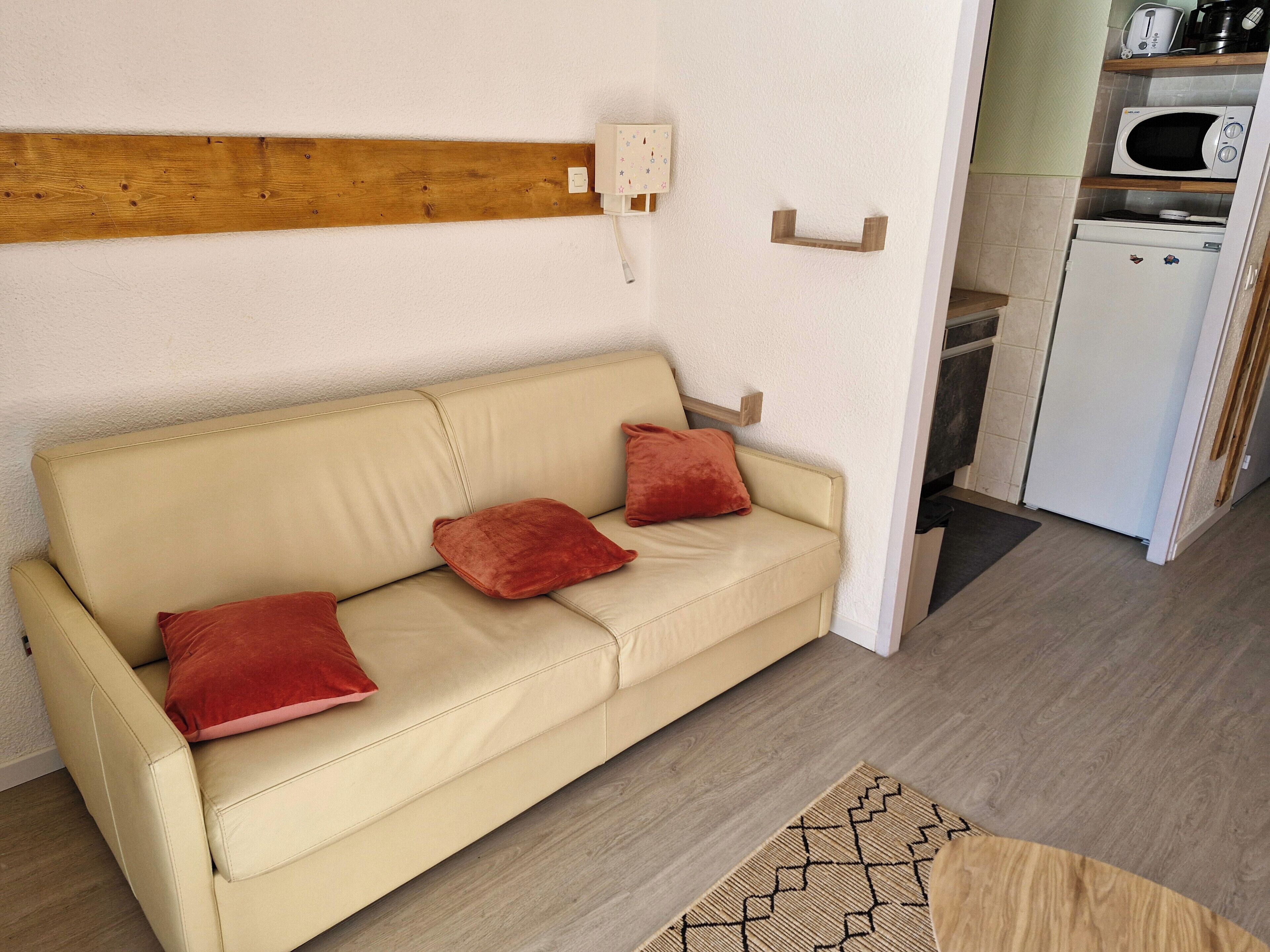 Un canapé beige avec des coussins rouges, dans une pièce ornée d’une poutre en bois au mur, un micro-ondes blanc et un petit réfrigérateur.