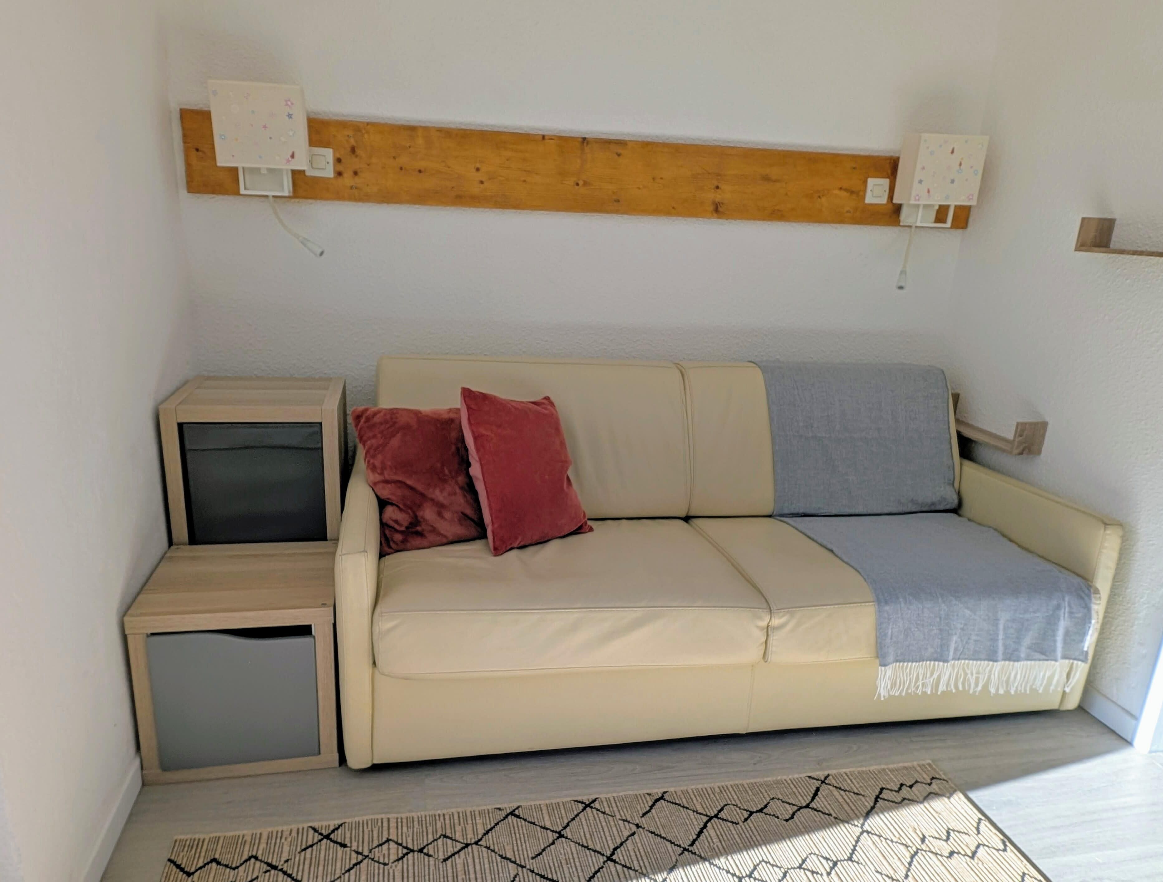 A small, cozy room with a beige sofa, two pillows, a small wooden side table, and a patterned rug.