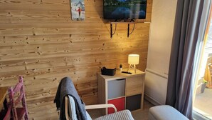 Une pièce chaleureuse avec un mur en bois, un téléviseur fixé au-dessus d’une petite table sur laquelle se trouve une lampe, une chaise et un banc.