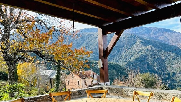 Une table ronde en bois, entourée de six chaises, est placée sous une pergola en bois. De là, on peut admirer un paysage montagneux aux couleurs automnales, avec un bâtiment rustique en arrière-plan.