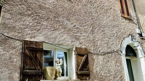 Un bâtiment rustique doté d’une façade à la texture particulière, de volets en bois et d’une petite fenêtre ornée d’une statue.
