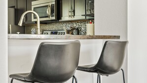 Una cucina moderna con mobili scuri, elettrodomestici in acciaio inossidabile e un piano di lavoro bianco. Due sgabelli neri sono posti di fronte al bancone.