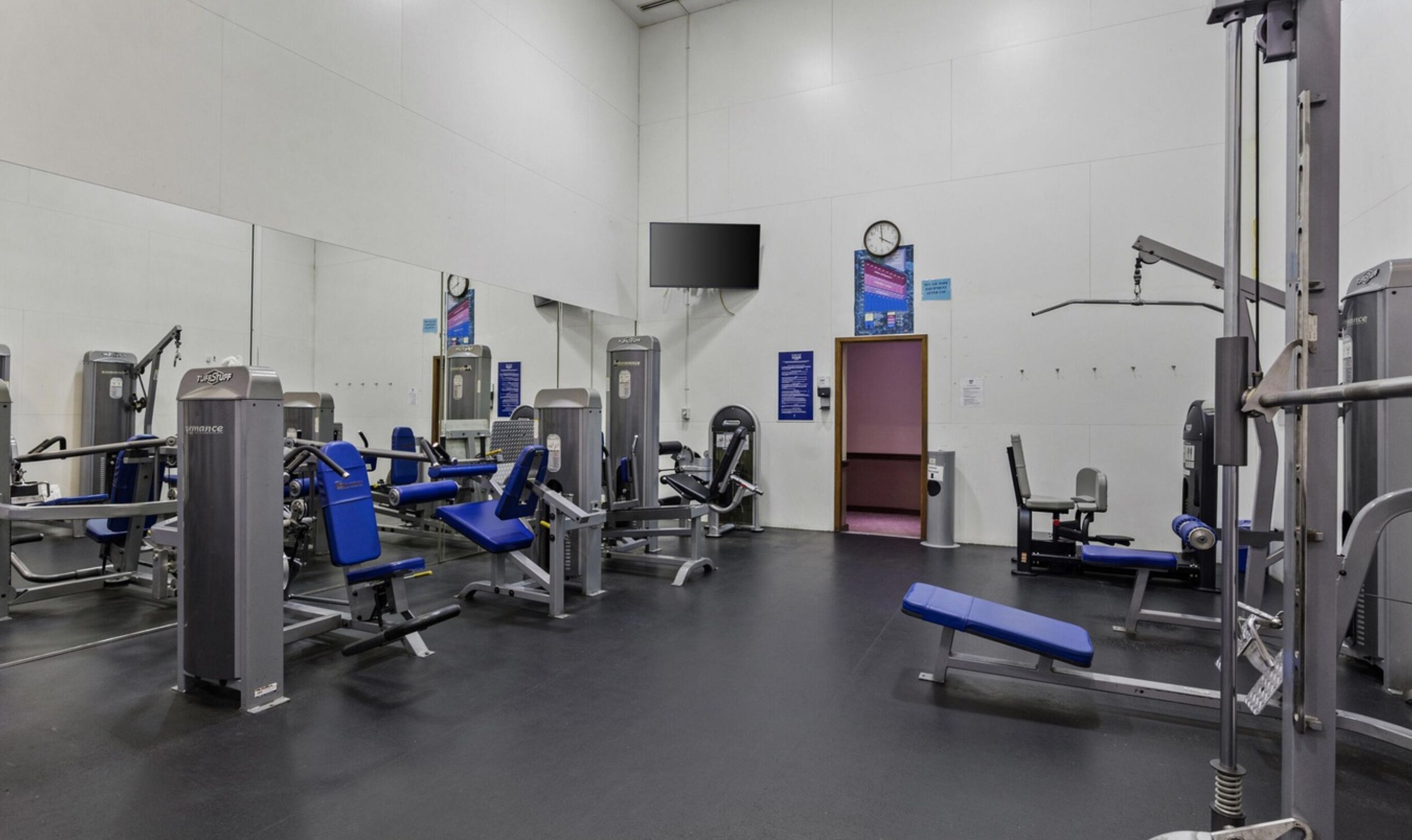 Une salle de sport équipée de divers appareils d’exercice, d’un téléviseur fixé au mur et d’une horloge.