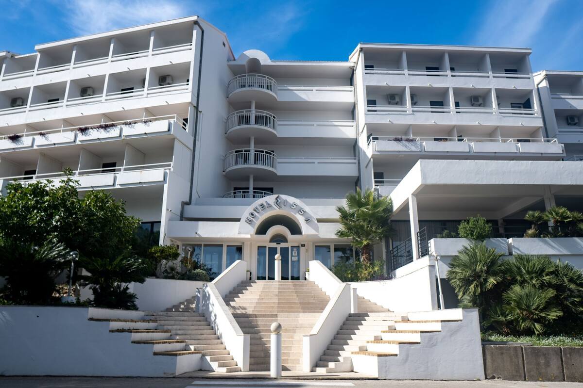 Um hotel moderno com entrada central, várias varandas e uma escadaria que leva à entrada.